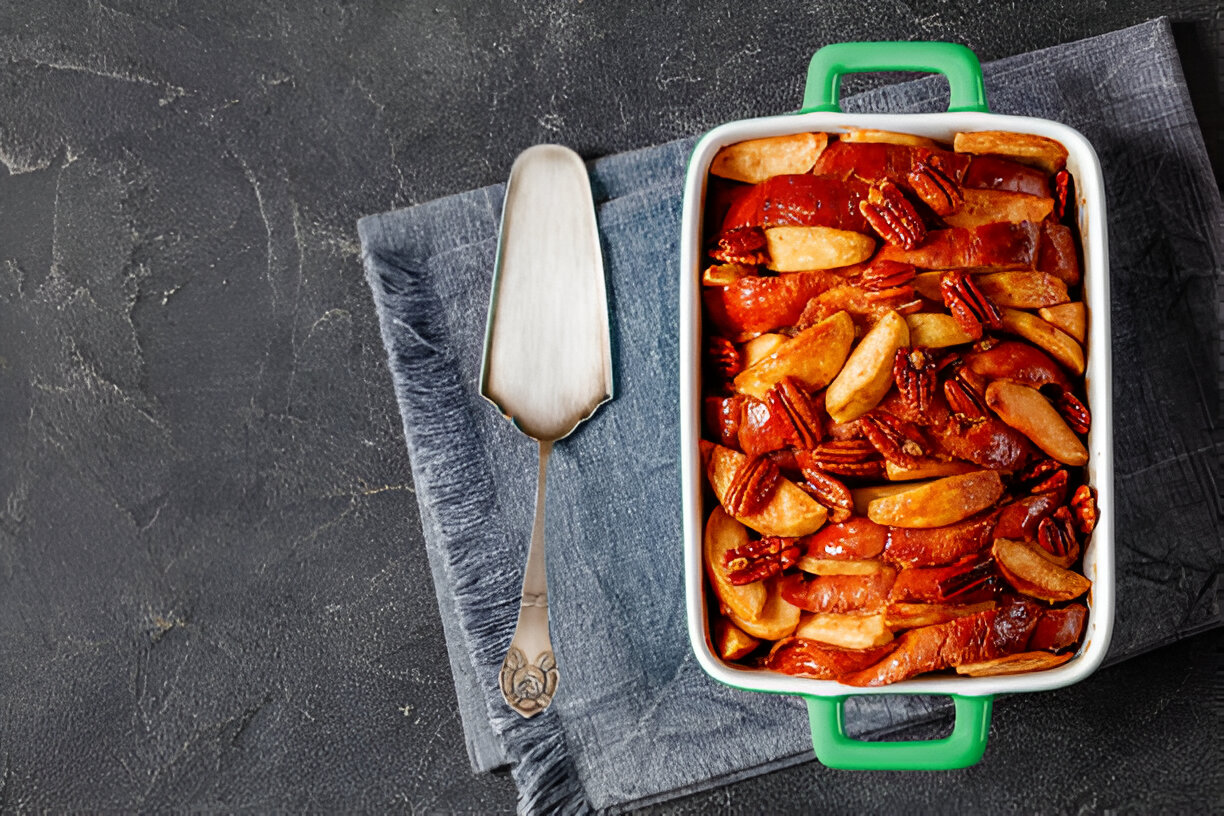 A baking dish filled with sliced apples and pecans, placed on a dark textured surface.