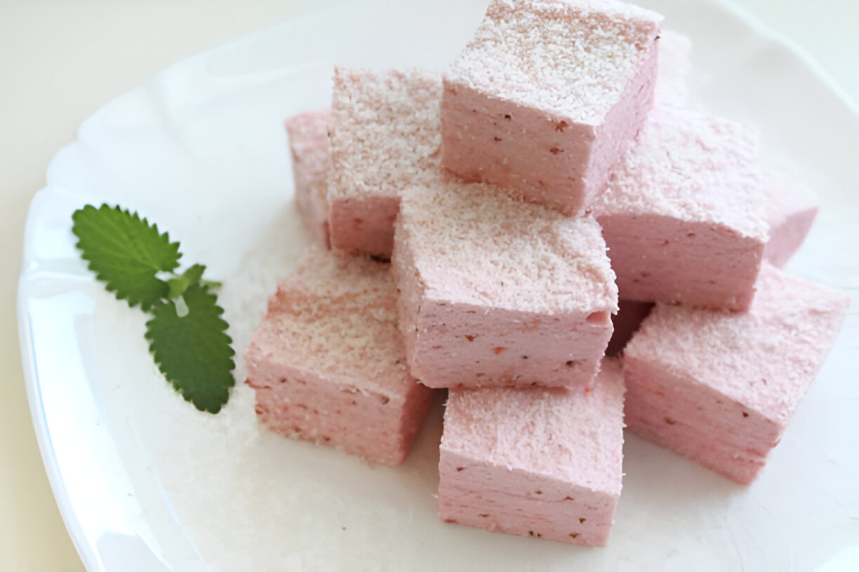 Square pieces of pink marshmallow with coconut flakes on a white plate, garnished with mint leaves.