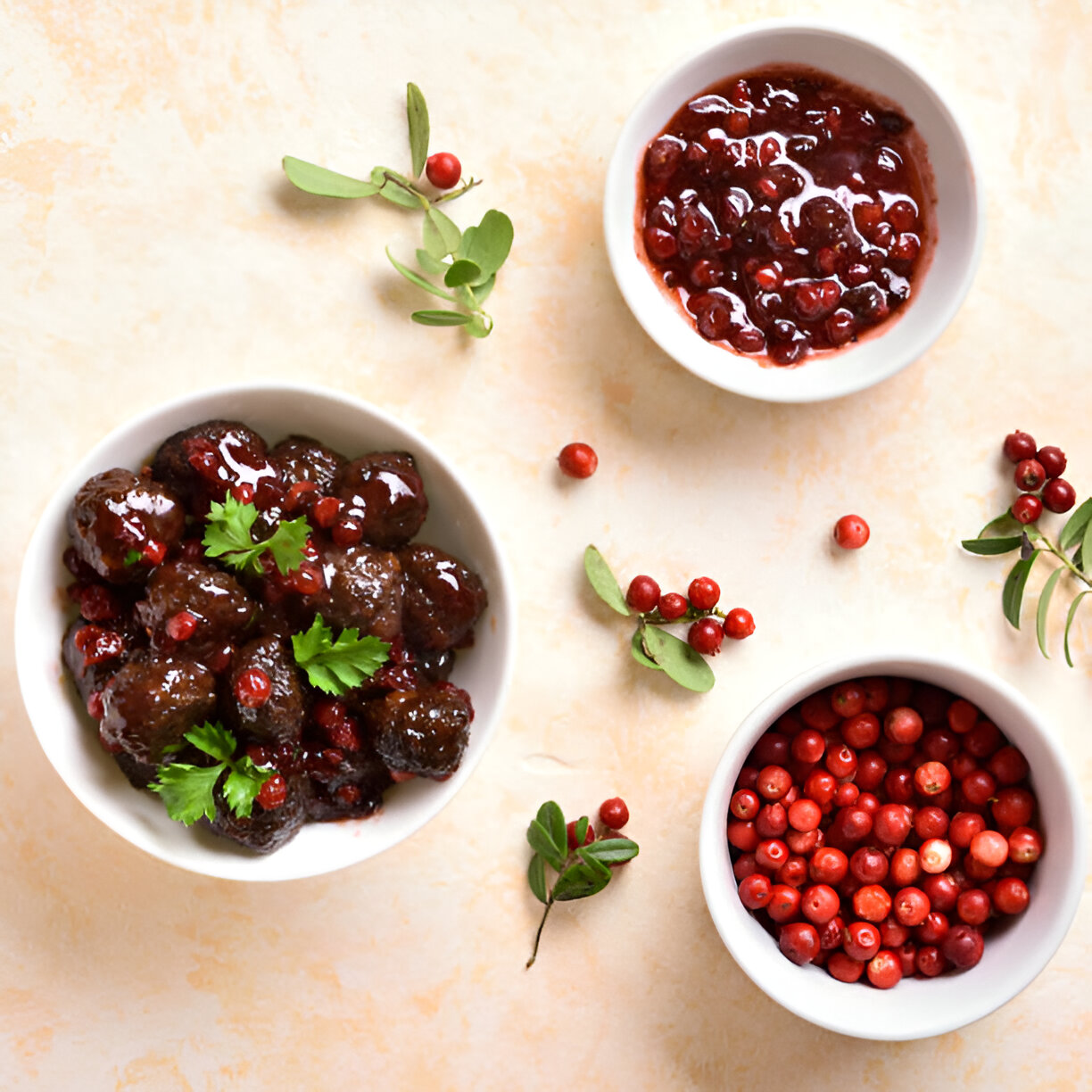 Bowls of dark, glossy meatballs garnished with cilantro, accompanied by red berry sauces and fresh red berries on a light background.