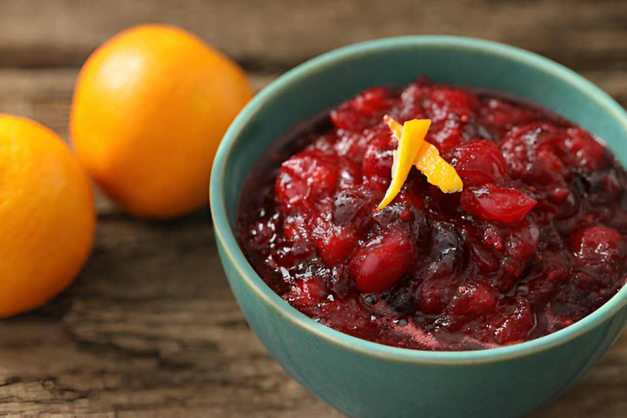 A teal bowl filled with cranberry sauce topped with orange zest, with two oranges in the background on a wooden surface.