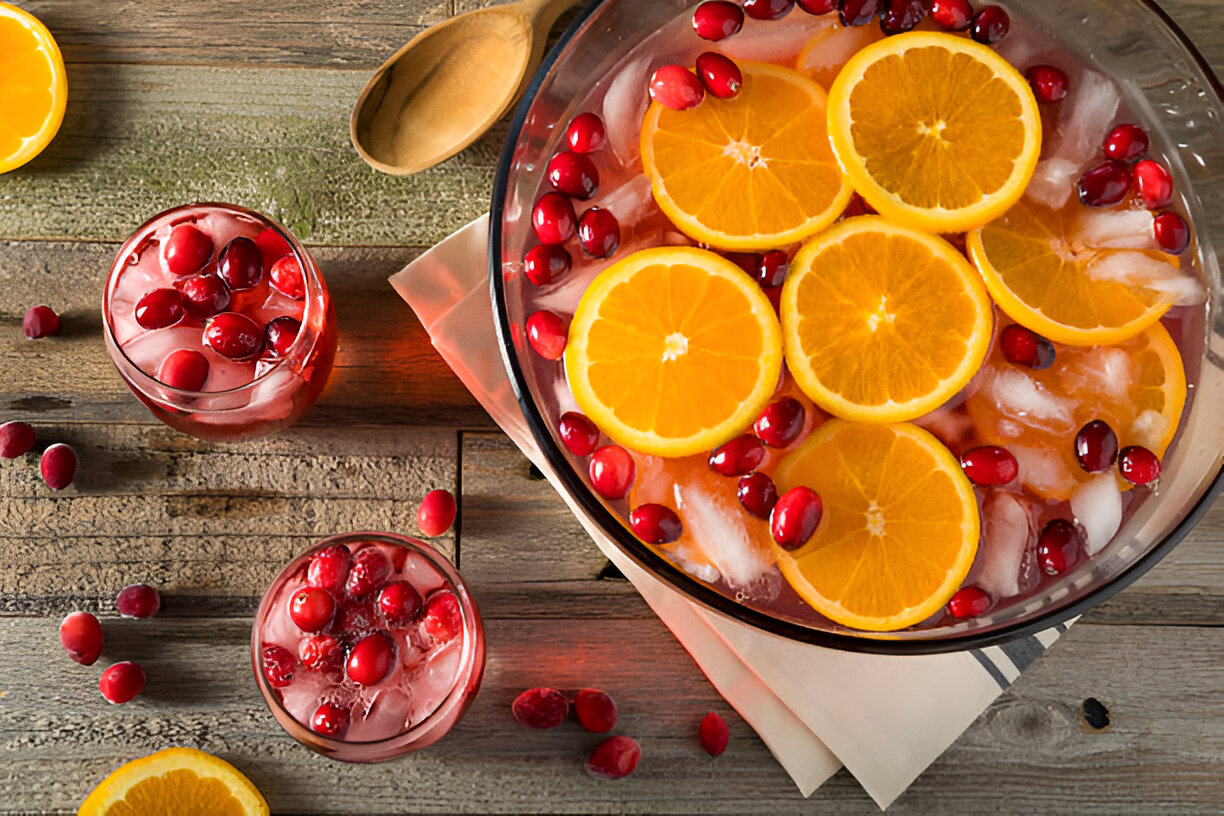 A punch bowl filled with sliced oranges, cranberries, and ice, alongside two glasses of cranberry drink on a wooden table.