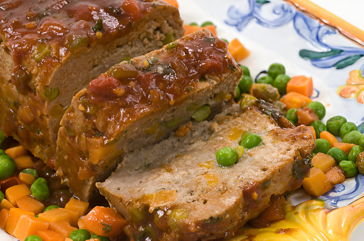A sliced meatloaf topped with tomato sauce, surrounded by peas and diced carrots on a decorative plate.