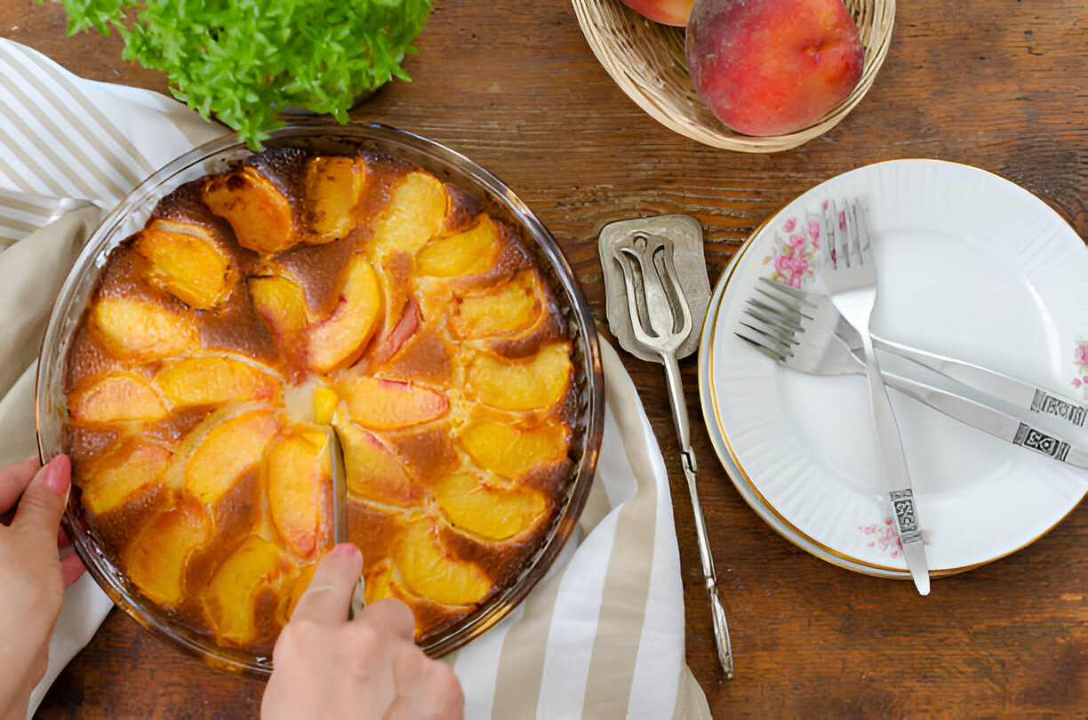 A freshly baked peach dessert being served with a knife on a wooden table decorated with a plant and plates.