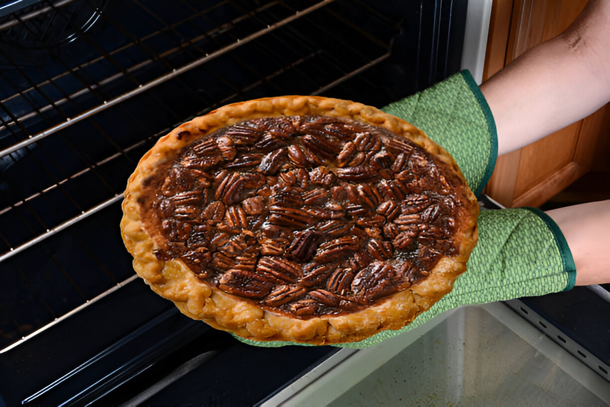 A person wearing green oven mitts is holding a freshly baked pecan pie from the oven.