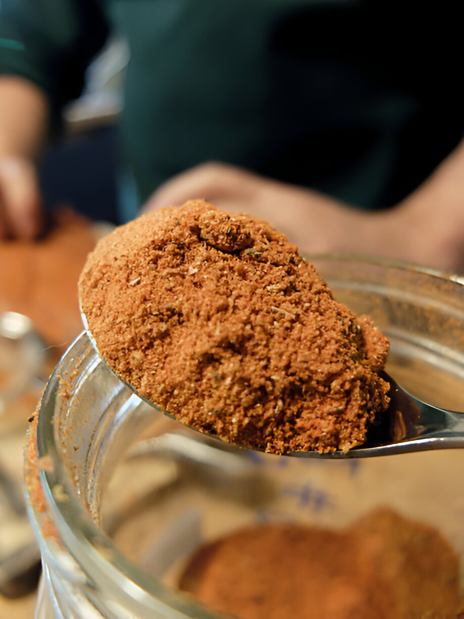 A spoon holding a heap of brown spice taken from a glass jar, with a blurred background showing a hand and other kitchen items.