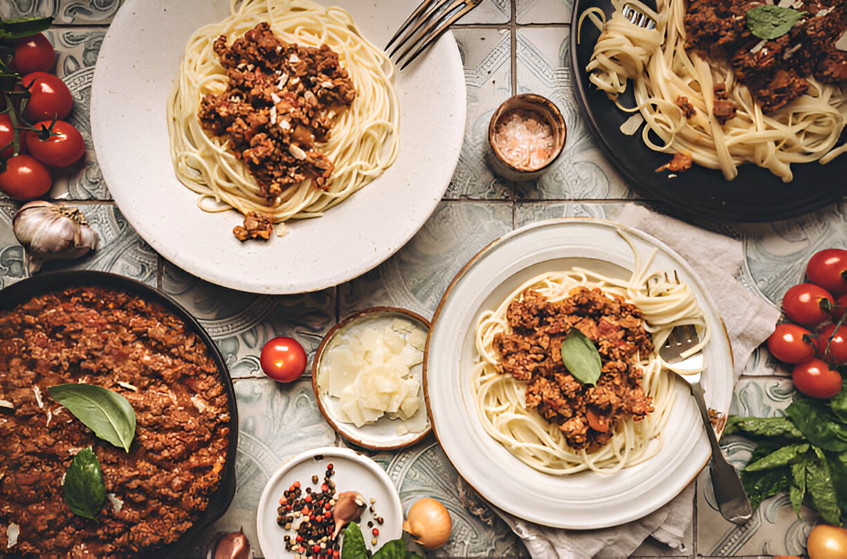 A beautifully arranged plate of spaghetti topped with a rich meat sauce, surrounded by fresh ingredients including cherry tomatoes, garlic, and herbs.