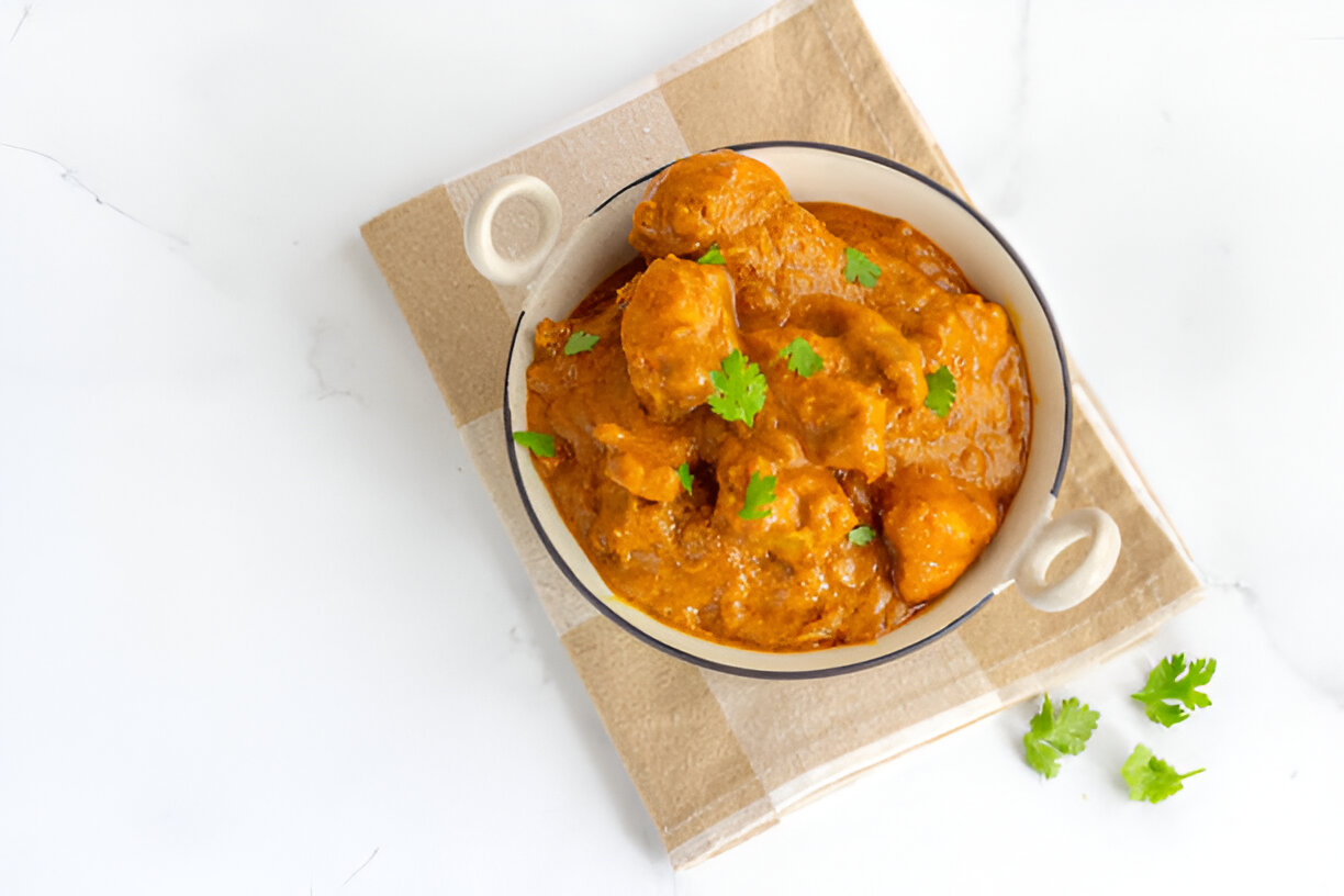 A bowl of rich curry with pieces of chicken garnished with fresh cilantro, placed on a beige cloth.