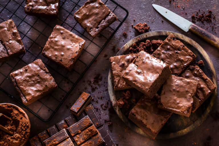 A wooden plate filled with delicious chocolate brownies next to a cooling rack with more brownies and chocolate bars.