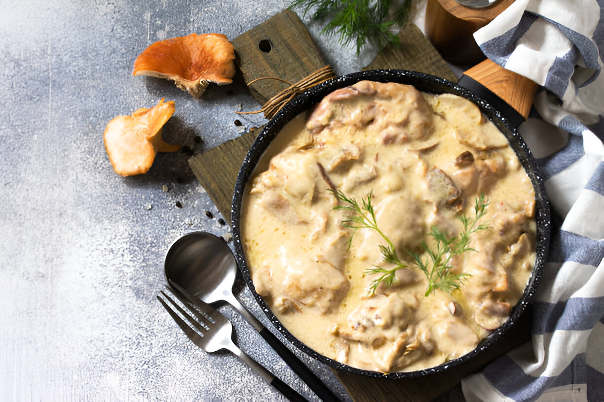 A delicious creamy mushroom dish served in a black skillet on a wooden cutting board, garnished with fresh dill, alongside a striped napkin and utensils.