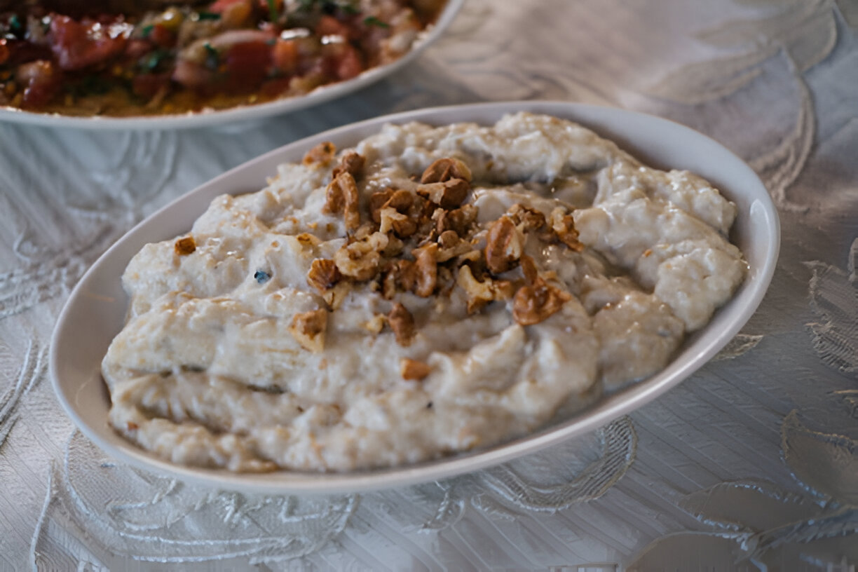 A creamy dish garnished with crushed nuts served in a white bowl, with a side dish of vibrant vegetables in the background.