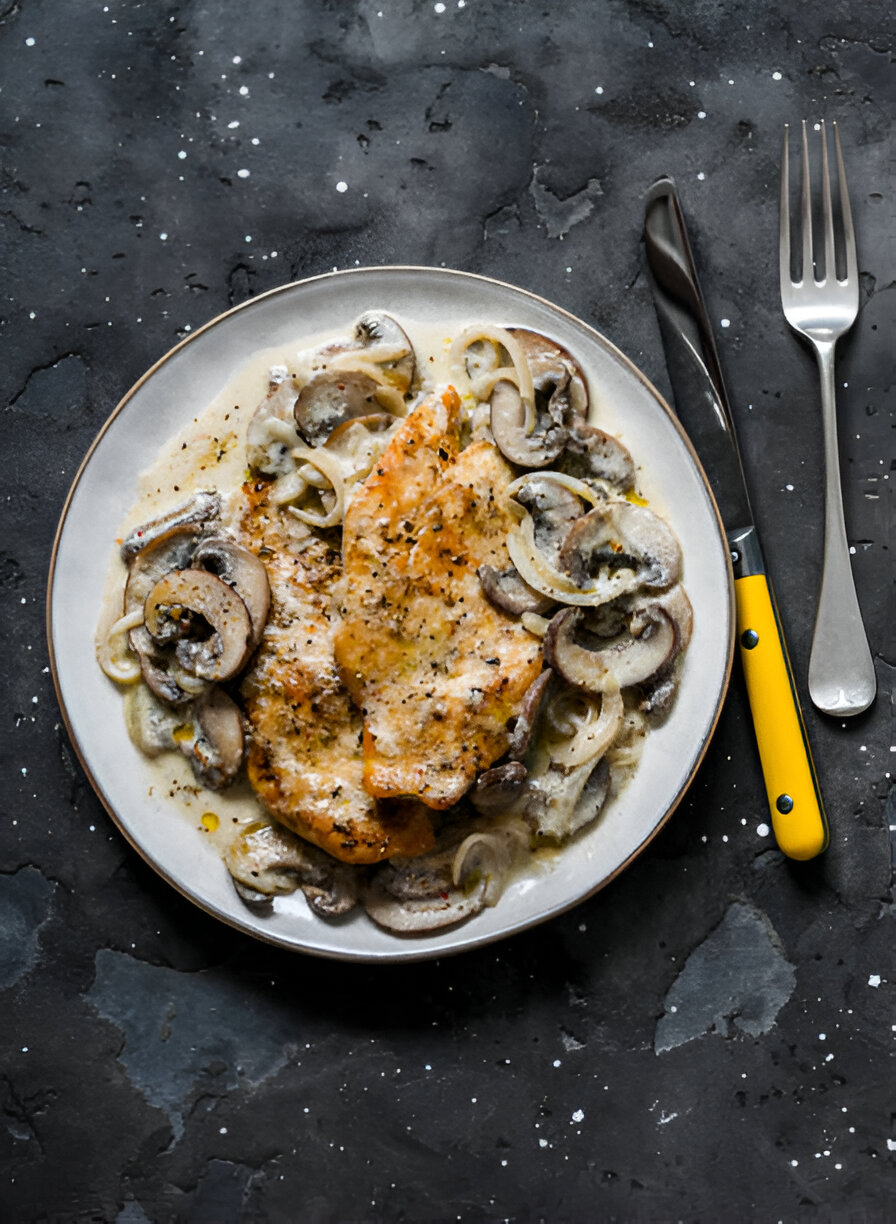 A plate of cooked fish fillets served with sautéed mushrooms and onions, set against a dark textured background.