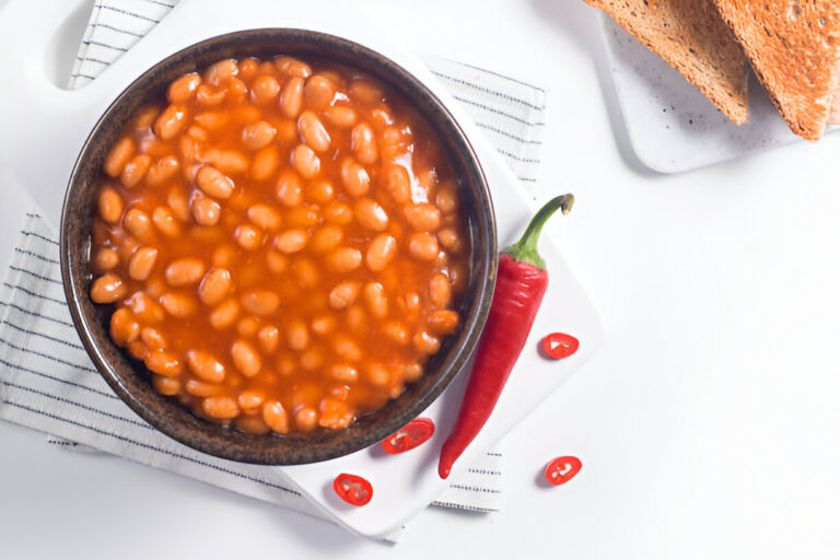 A bowl of baked beans in a rich tomato sauce surrounded by red chili peppers and slices of toasted bread.