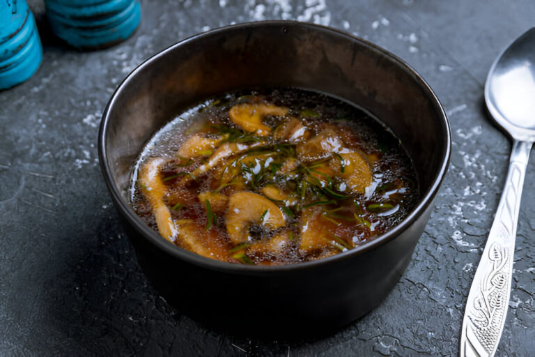 A bowl of flavorful broth with mushrooms and green herbs, placed on a textured, dark surface.