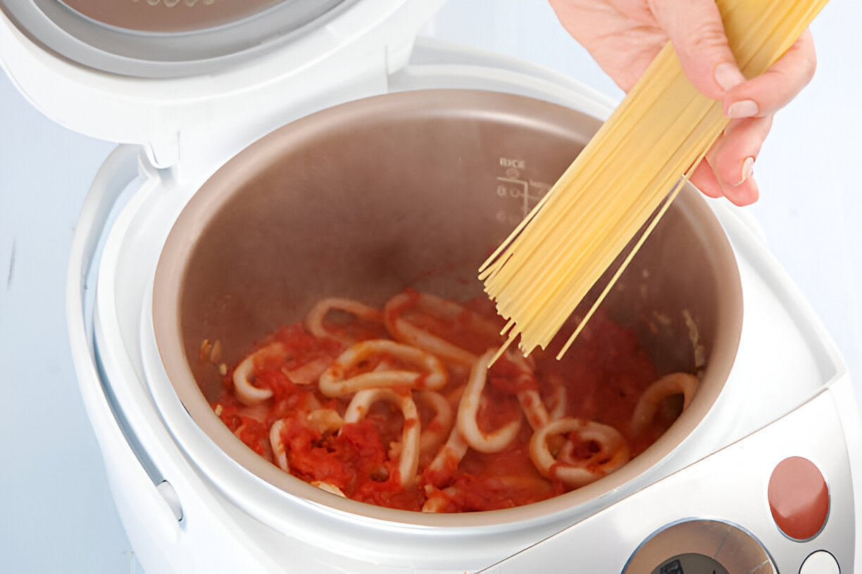 A person's hand holding uncooked spaghetti over a pot of tomato sauce with pasta cooking inside a rice cooker.