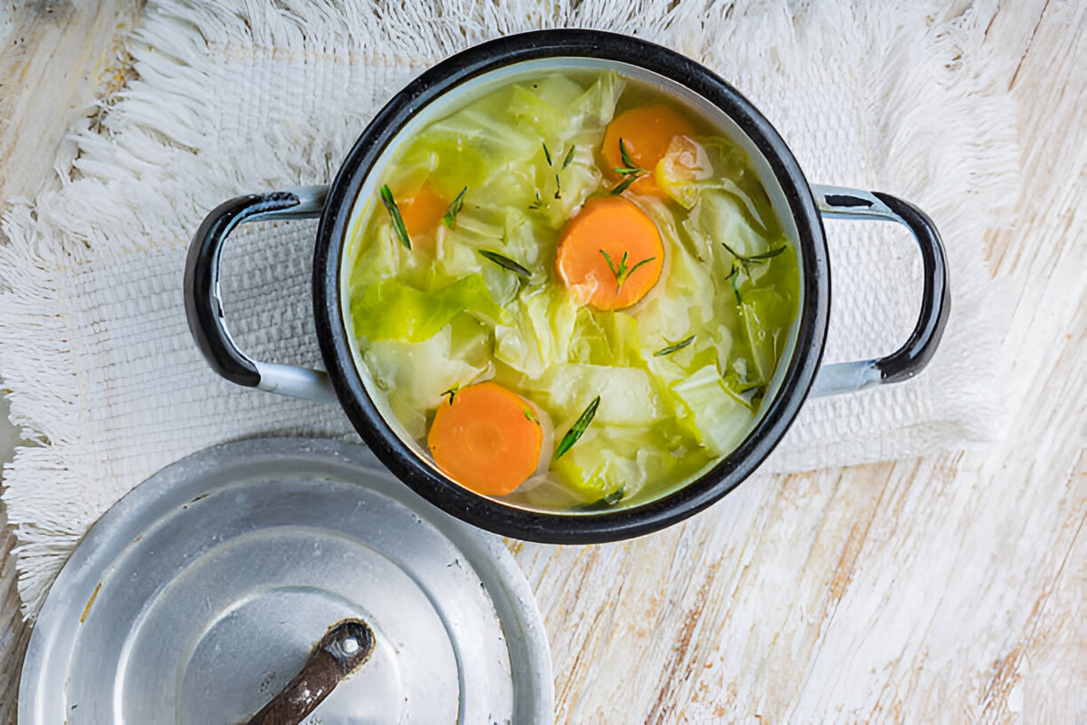 A pot of vegetable soup containing cabbage, carrots, and herbs on a textured surface.