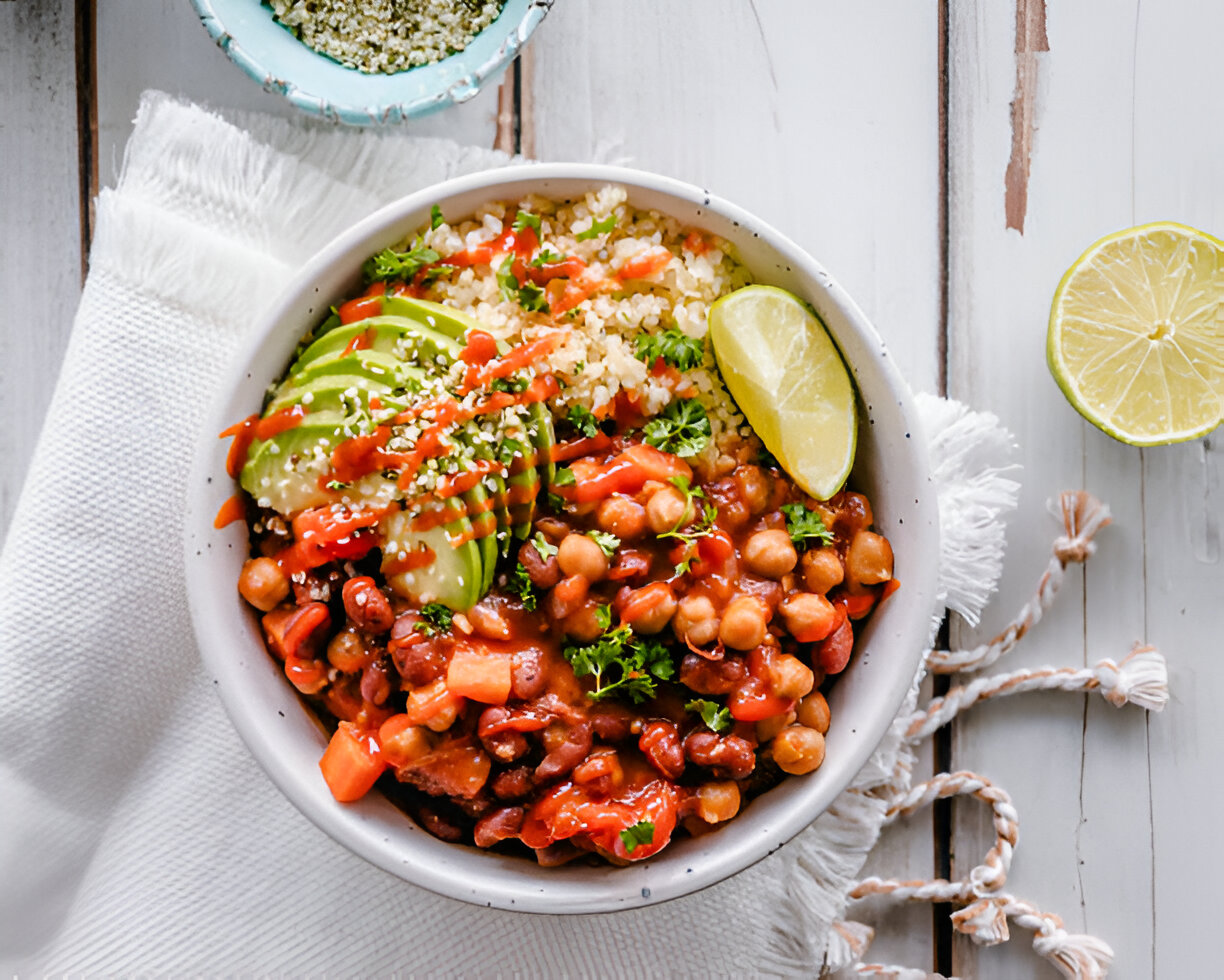 A colorful bowl of chili with chickpeas, beans, avocado slices, and quinoa, garnished with lime and sesame seeds.