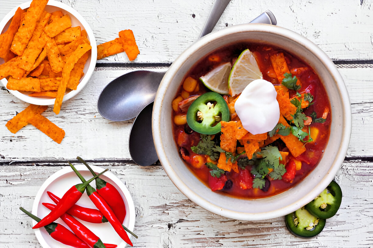 A bowl of vibrant chili topped with sour cream, fresh cilantro, jalapeño slices, and tortilla strips, accompanied by a side of red chili peppers and tortilla chips.