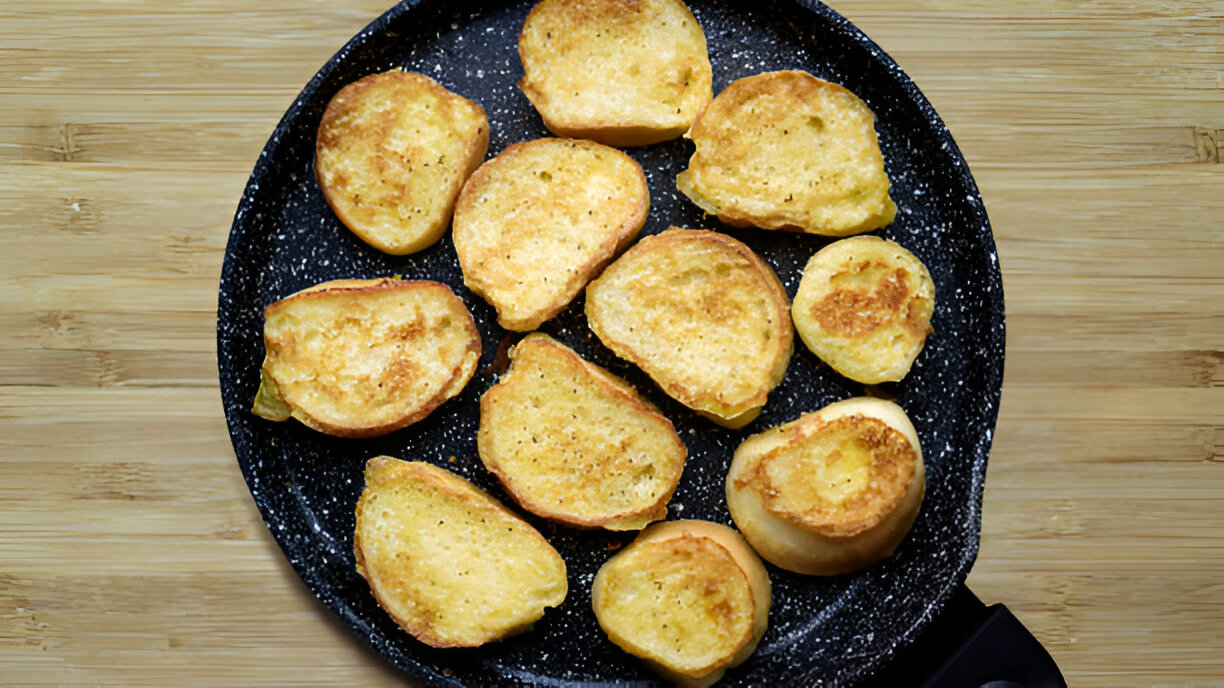 A skillet filled with golden-brown fried bread slices on a wooden surface.