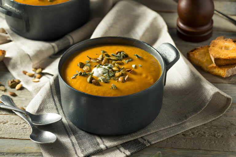 A bowl of creamy orange soup garnished with seeds and herbs, served on a textured fabric with spoons and toasted bread in the background.