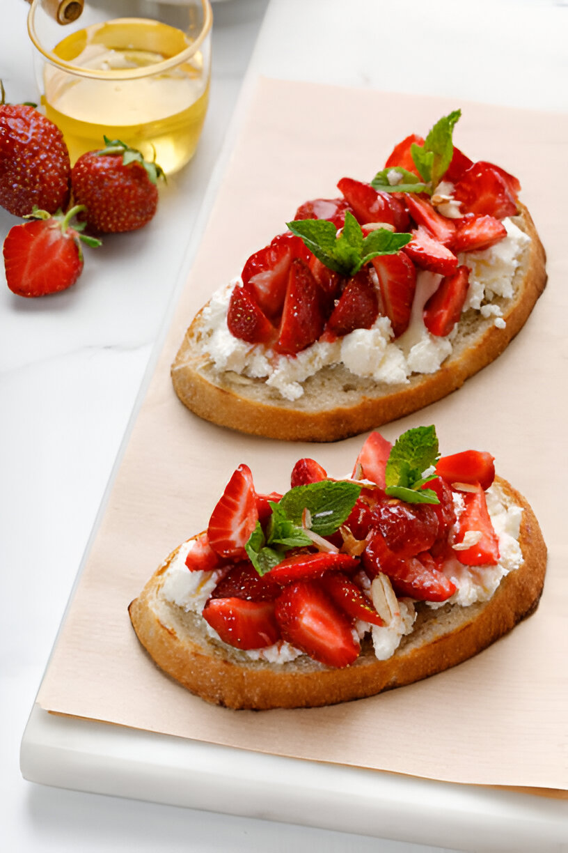 Two slices of bread topped with fresh strawberries, mint leaves, and cream cheese, with strawberries and olive oil in the background.