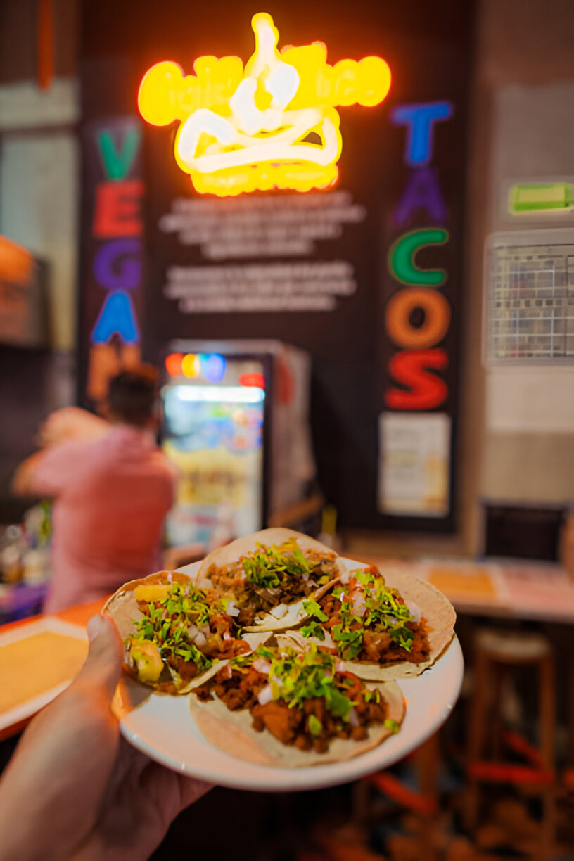 A hand holding a plate of three tacos topped with vegetables, with a colorful restaurant background and a neon sign.