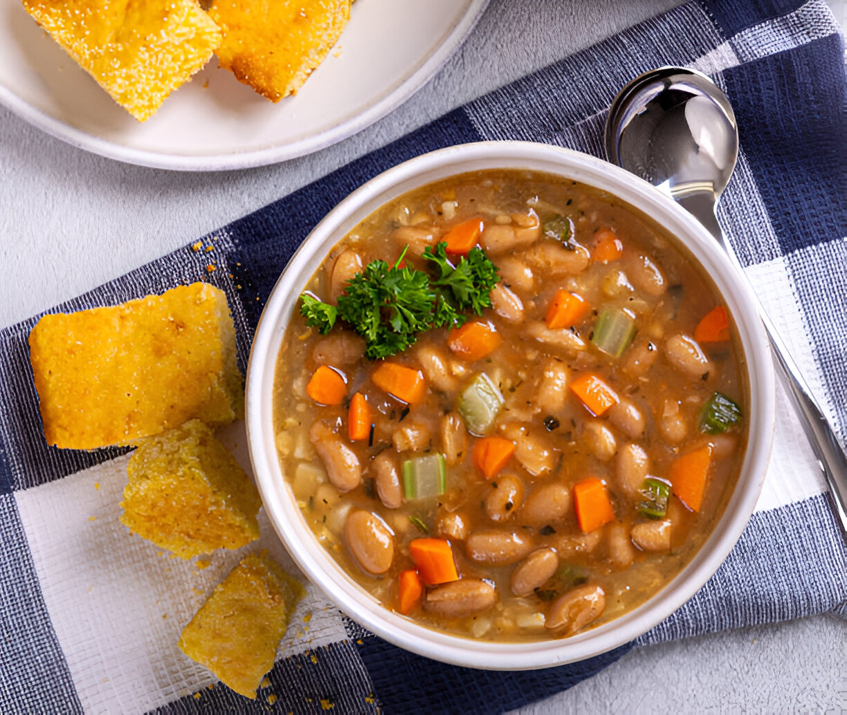 A bowl of hearty bean soup with diced vegetables, garnished with parsley, served alongside cornbread pieces.