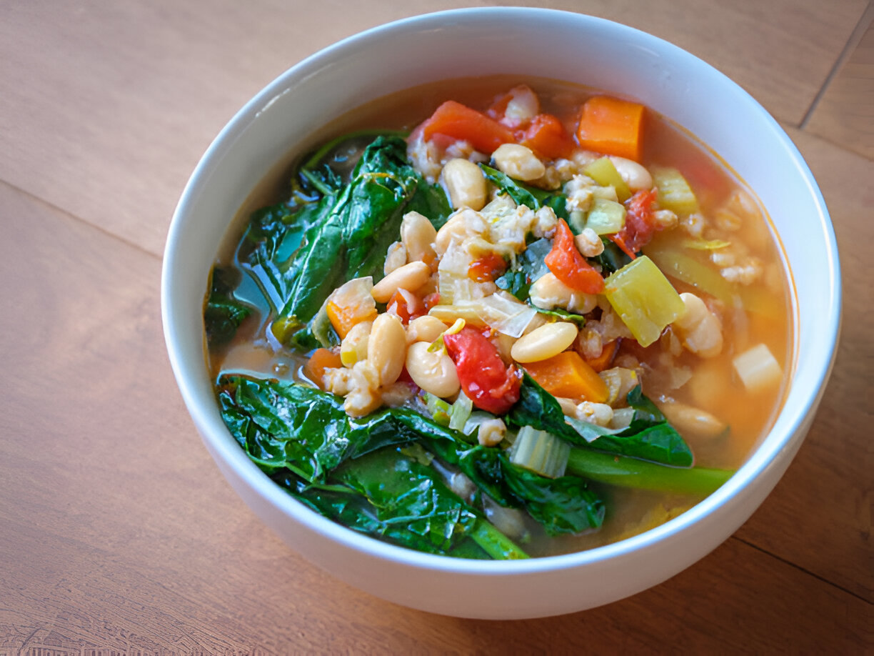 A bowl of vegetable soup with beans and greens sitting on a wooden surface.