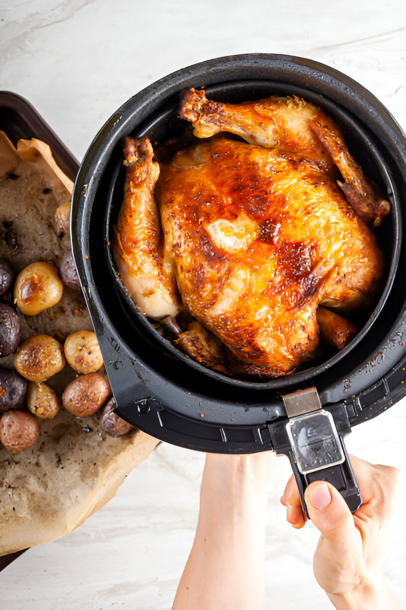 A freshly cooked whole chicken being lifted from an air fryer alongside a tray of roasted multi-colored potatoes.