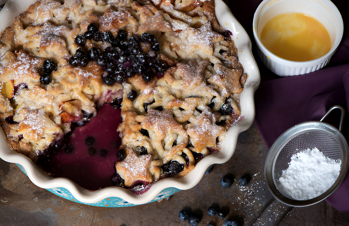 Homemade blueberry dessert in a pie dish with a piece cut out, surrounded by blueberries and powdered sugar.