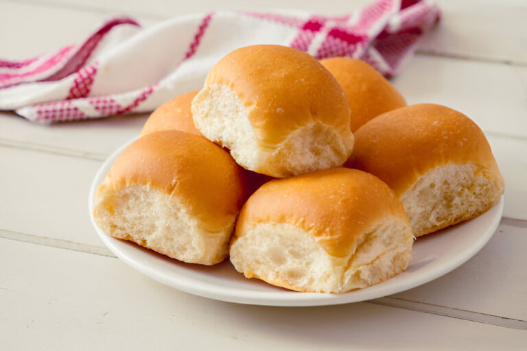 A plate of soft, golden dinner rolls stacked together, with a patterned cloth draped in the background.