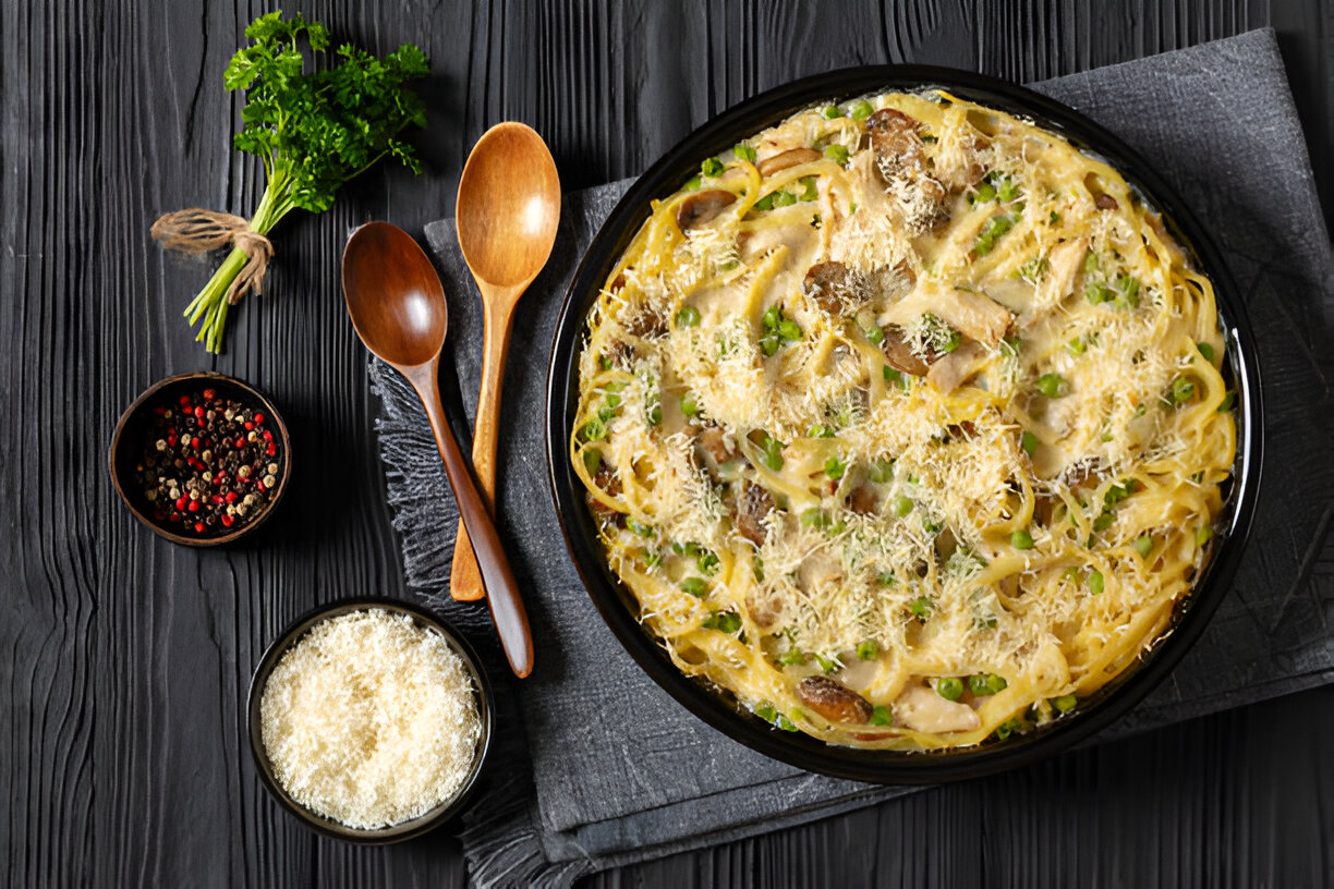 A delicious bowl of creamy pasta with green peas and mushrooms, topped with grated cheese, accompanied by wooden spoons and a bowl of peppercorns.