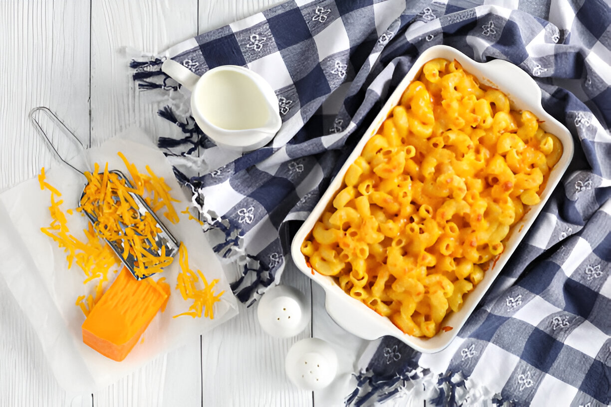 Creamy macaroni and cheese in a white dish with shredded cheese, milk, and a checkered napkin