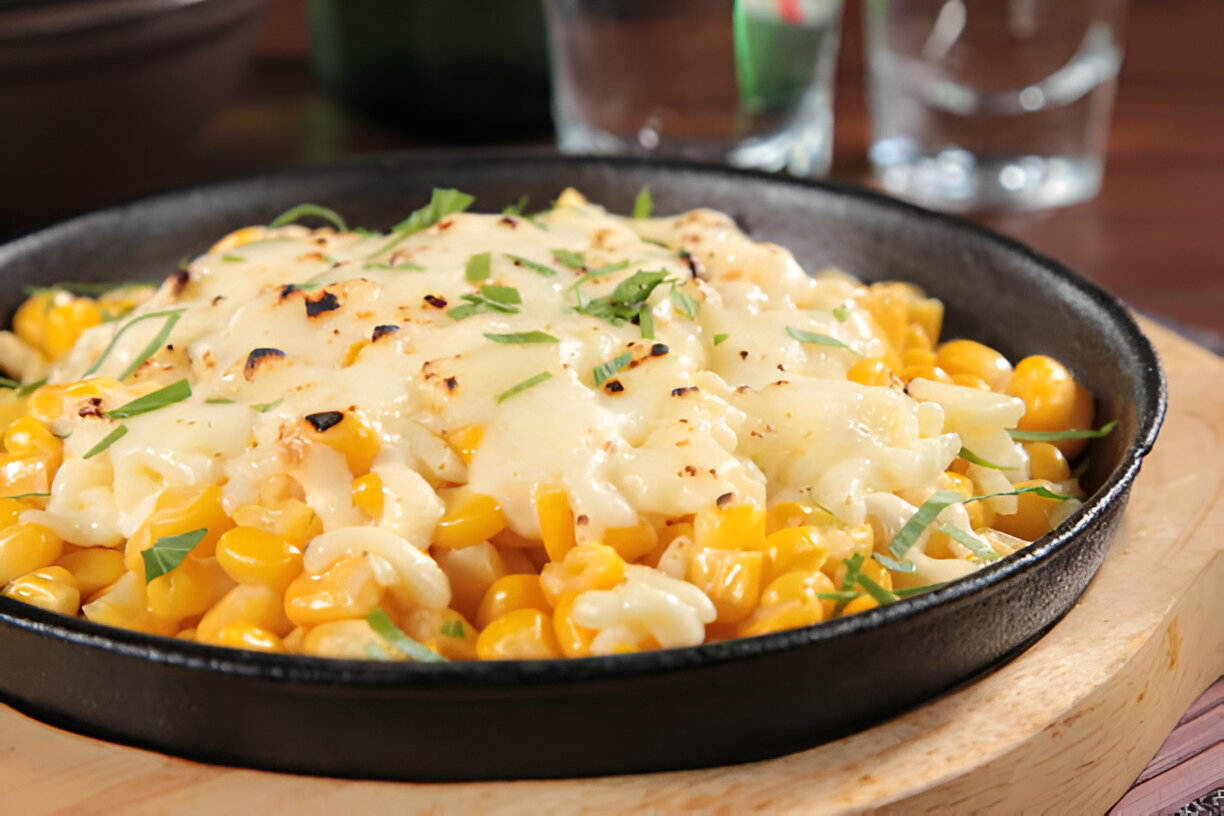 A close-up view of cheesy corn pasta served in a black skillet, topped with green herbs.