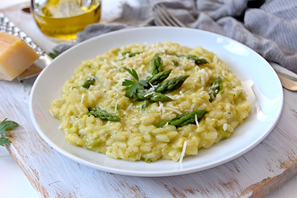 A plate of creamy asparagus risotto garnished with fresh asparagus tips and parsley, next to a piece of cheese and olive oil
