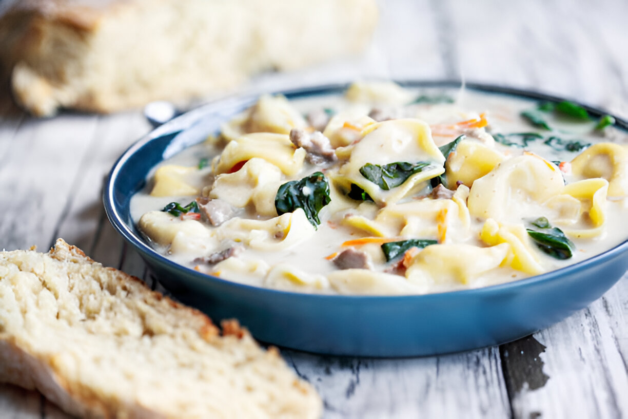 A bowl of creamy tortellini soup with spinach, served alongside a slice of bread.