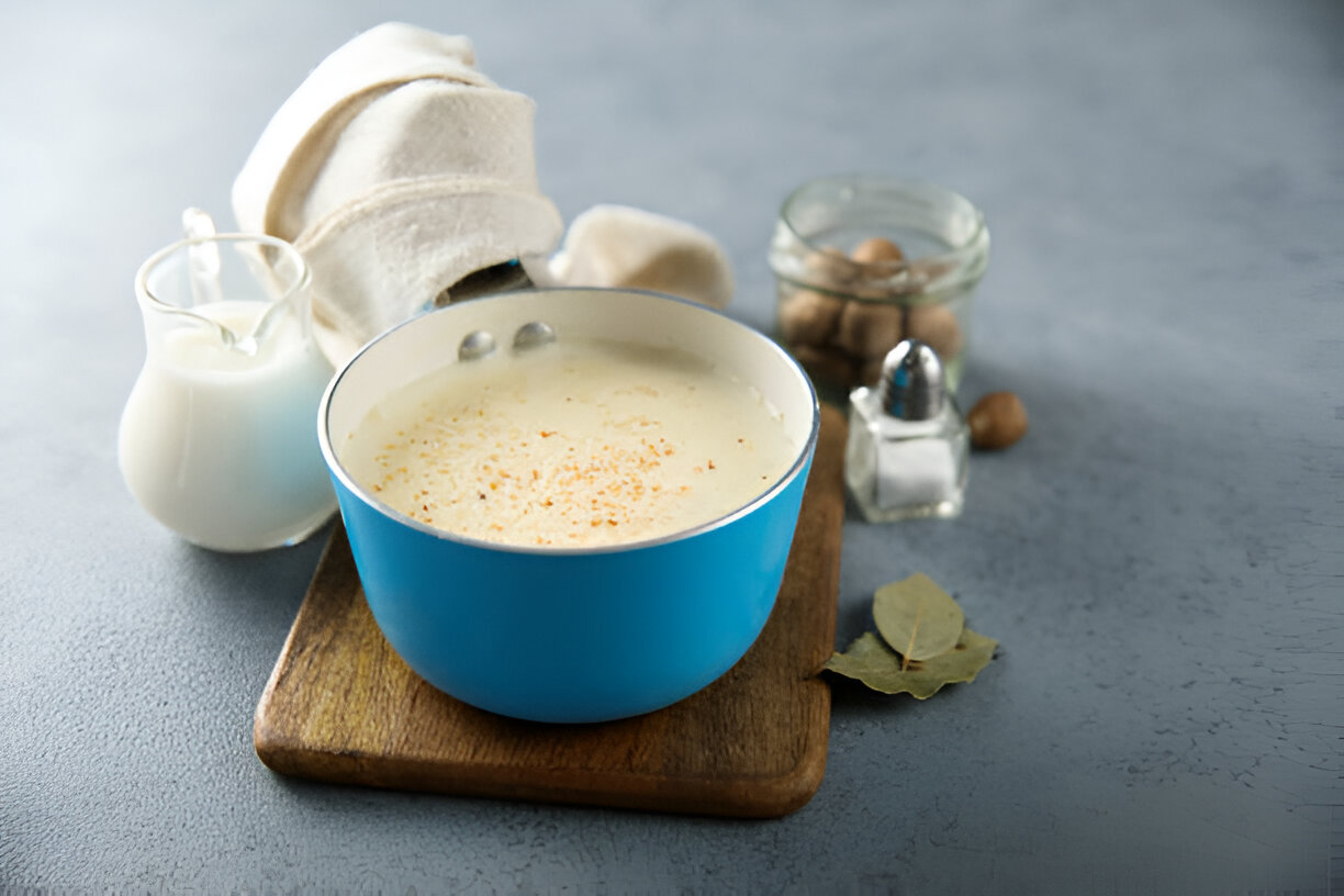 A blue pot filled with creamy mixture and topped with spices, placed on a wooden cutting board. Accompanied by a glass of milk, a shaker of salt, and a jar of whole spices.