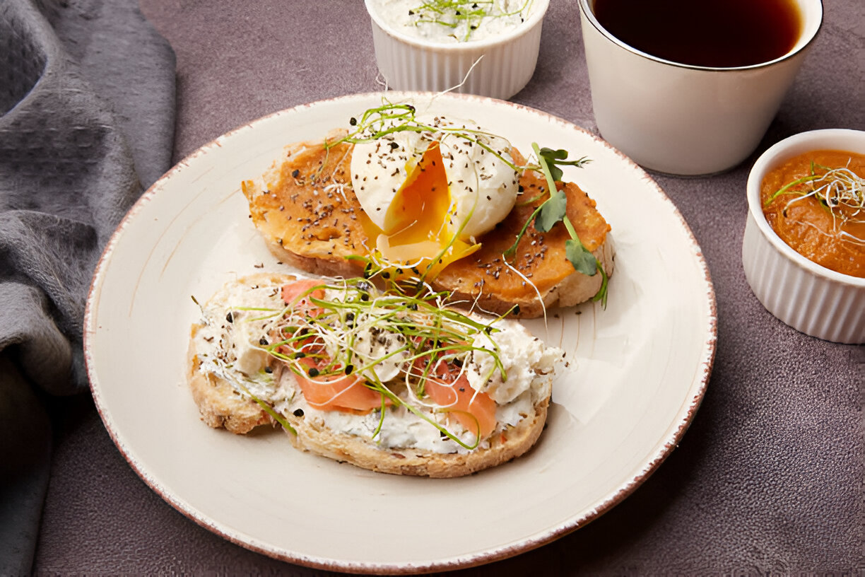 A plate of gourmet breakfast featuring bread topped with poached egg, smoked salmon, and microgreens, accompanied by small bowls of cream and soup.