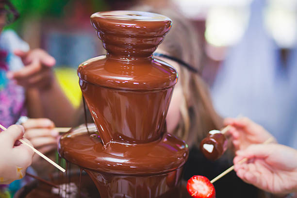 A chocolate fountain surrounded by people holding skewers with fruits and sweets.