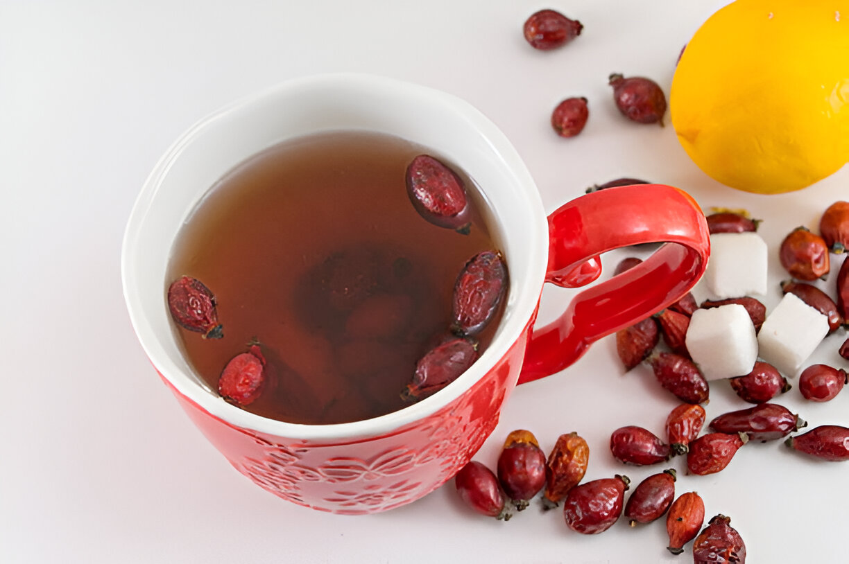 A red mug containing herbal tea with dried rose hips, surrounded by fresh lemon, sugar cubes, and more dried rose hips.