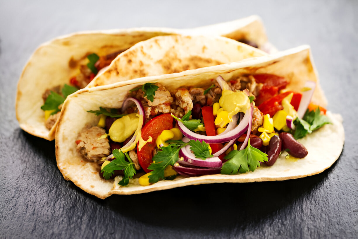 Two tacos filled with meat, beans, and fresh vegetables placed on a dark slate surface.