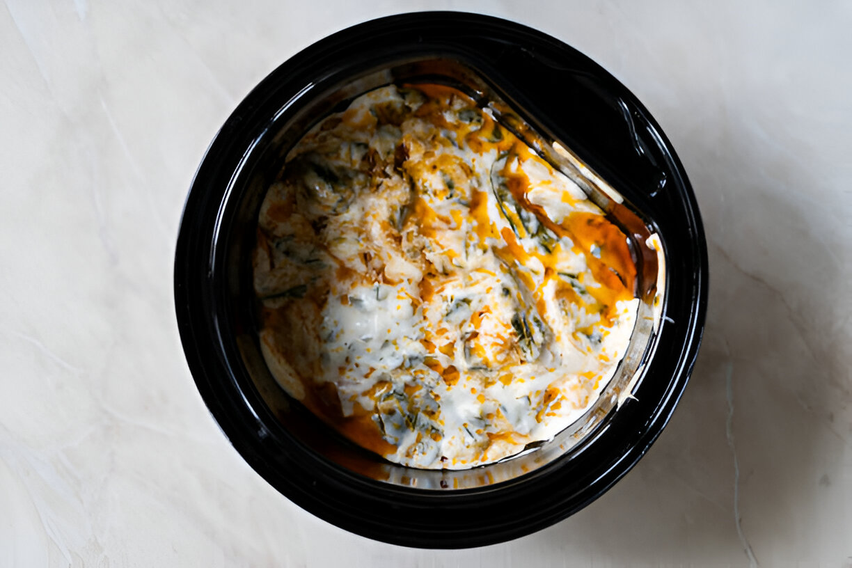 A close-up view of a slow cooker filled with a creamy and colorful dish, featuring layers of white and orange with some green elements.