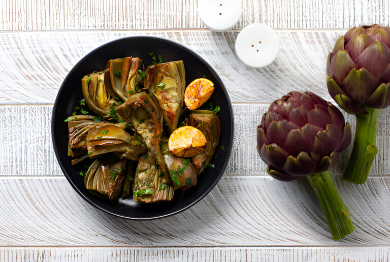 A black bowl filled with roasted artichokes and grilled lemon slices garnished with parsley on a wooden surface, surrounded by fresh artichokes and white candles.