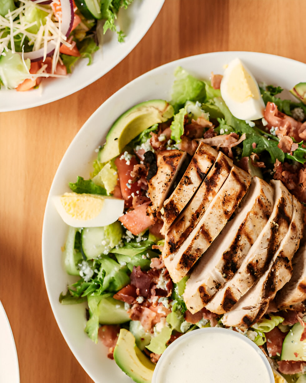 A delicious grilled chicken salad with sliced avocados, tomatoes, and boiled eggs, served with a side of ranch dressing.