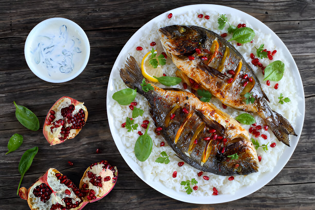 A beautifully plated dish of grilled fish served on a bed of rice, garnished with pomegranate seeds and fresh herbs, alongside a bowl of yogurt and halved pomegranates.