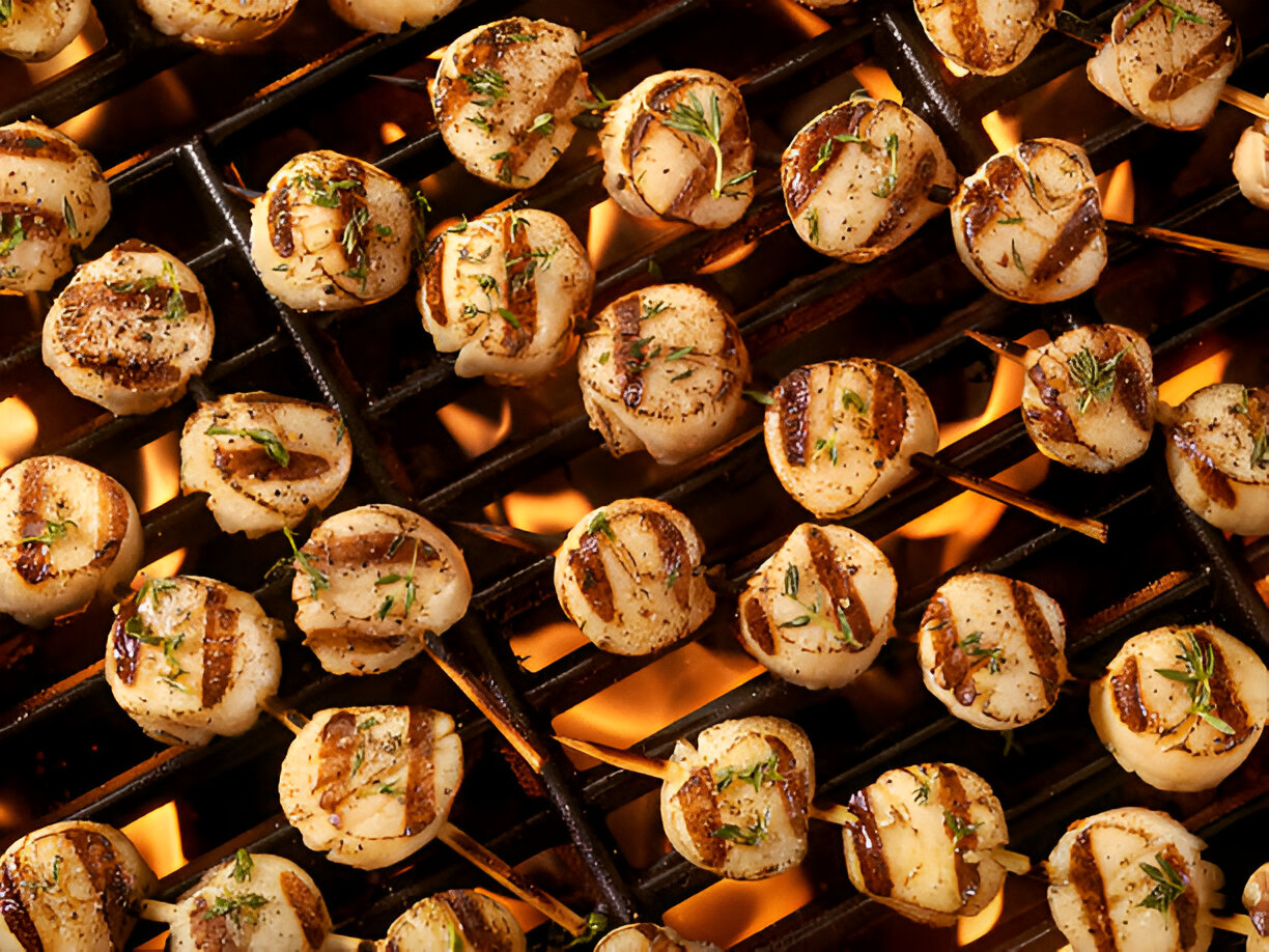 Grilled scallops skewered on sticks, garnished with herbs and placed on a BBQ grill over flames.