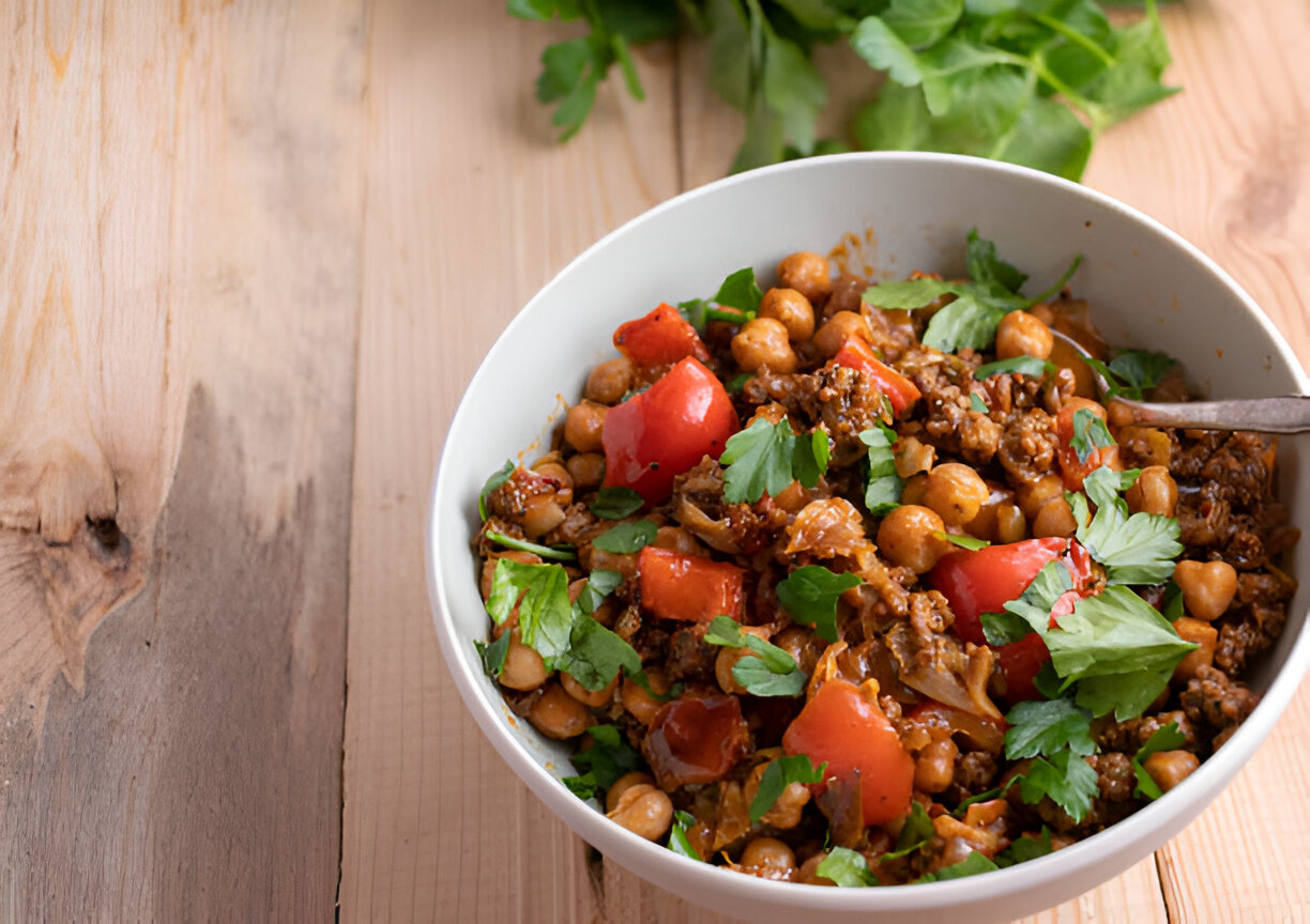 A bowl of chickpea and minced meat dish garnished with fresh parsley and red bell peppers on a wooden table.