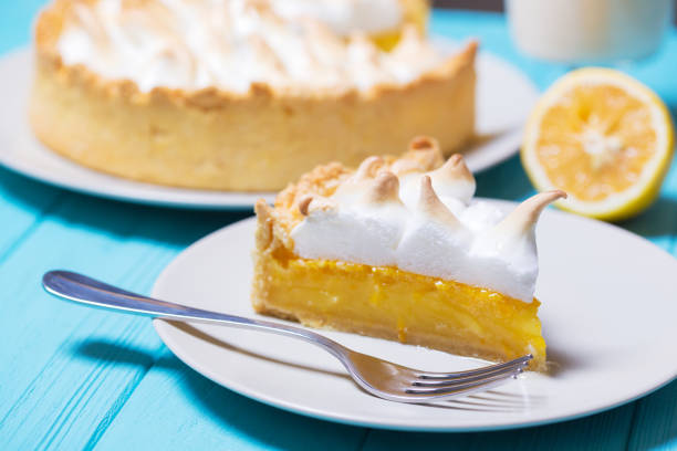 A slice of lemon meringue pie on a white plate with a fork, accompanied by a whole lemon and a whole pie in the background.