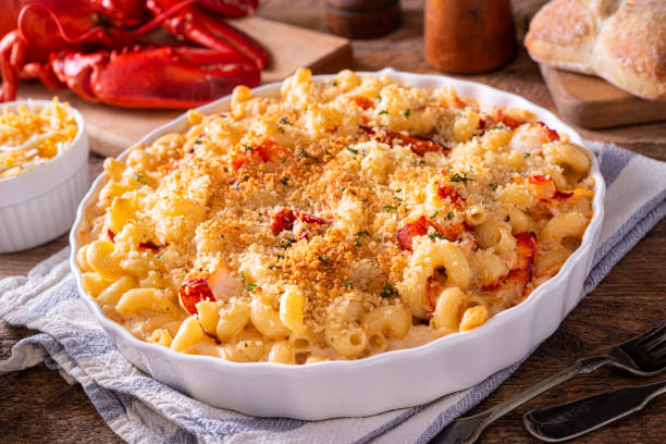 A creamy baked lobster mac and cheese in a white dish with a crunchy topping, served alongside lobster and bread.