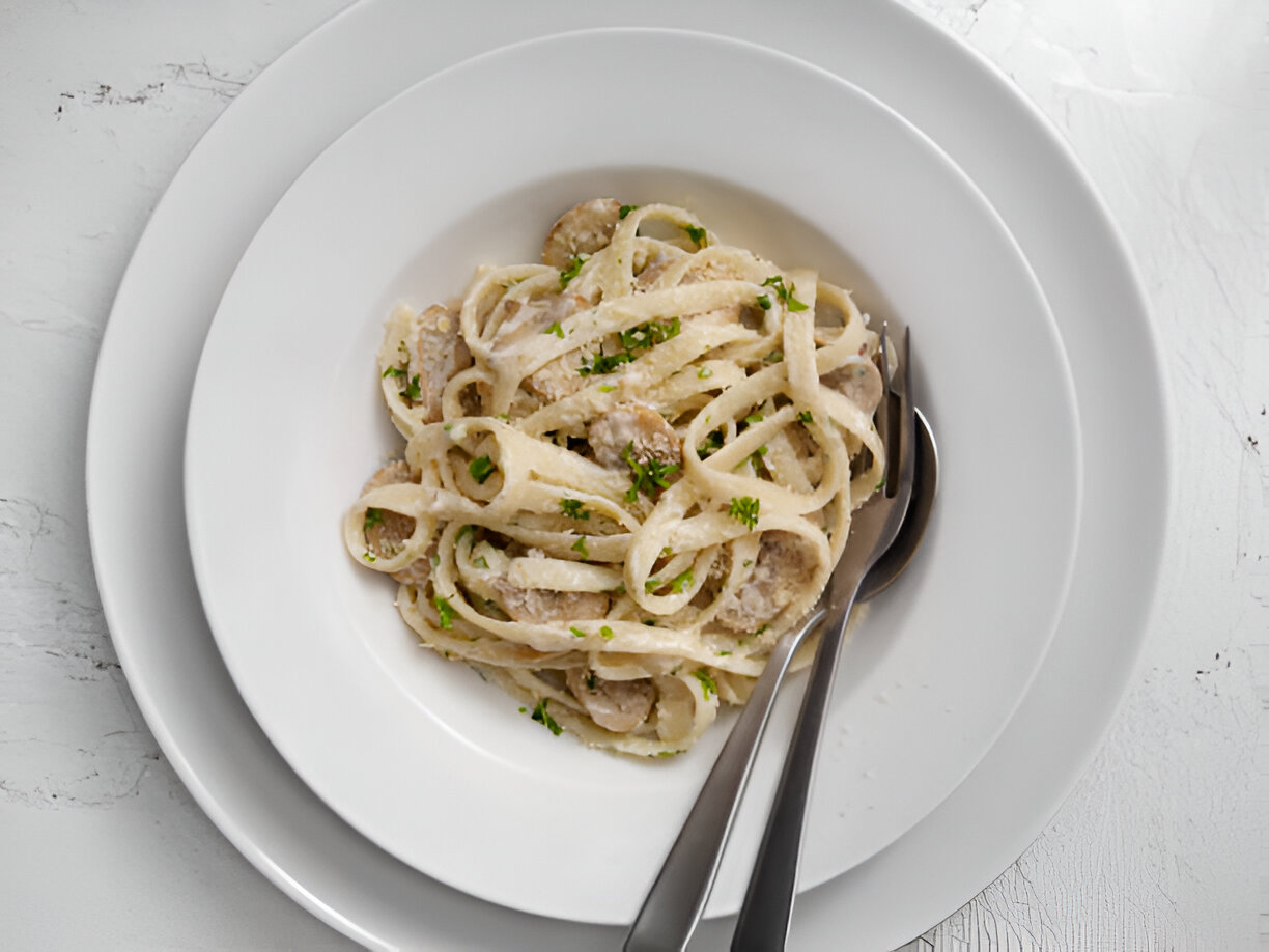 A delicious plate of fettuccine pasta served in a white bowl, garnished with parsley and accompanied by silver utensils.