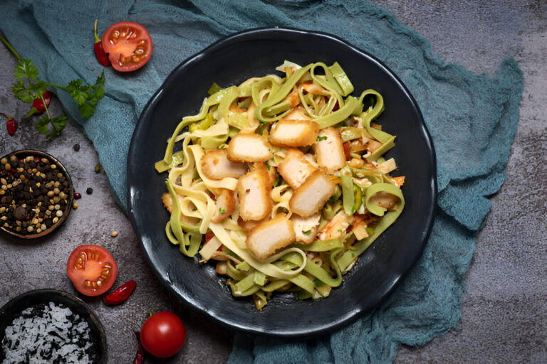 A black bowl filled with green and yellow fettuccine pasta topped with slices of breaded chicken, surrounded by fresh tomatoes and spices.
